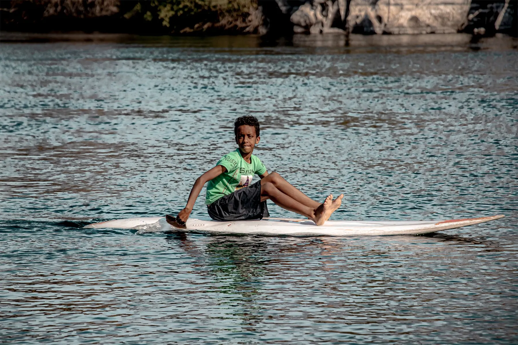A young paddler sitting on a board on the Nile at Aswan, with rocks and palms behind.