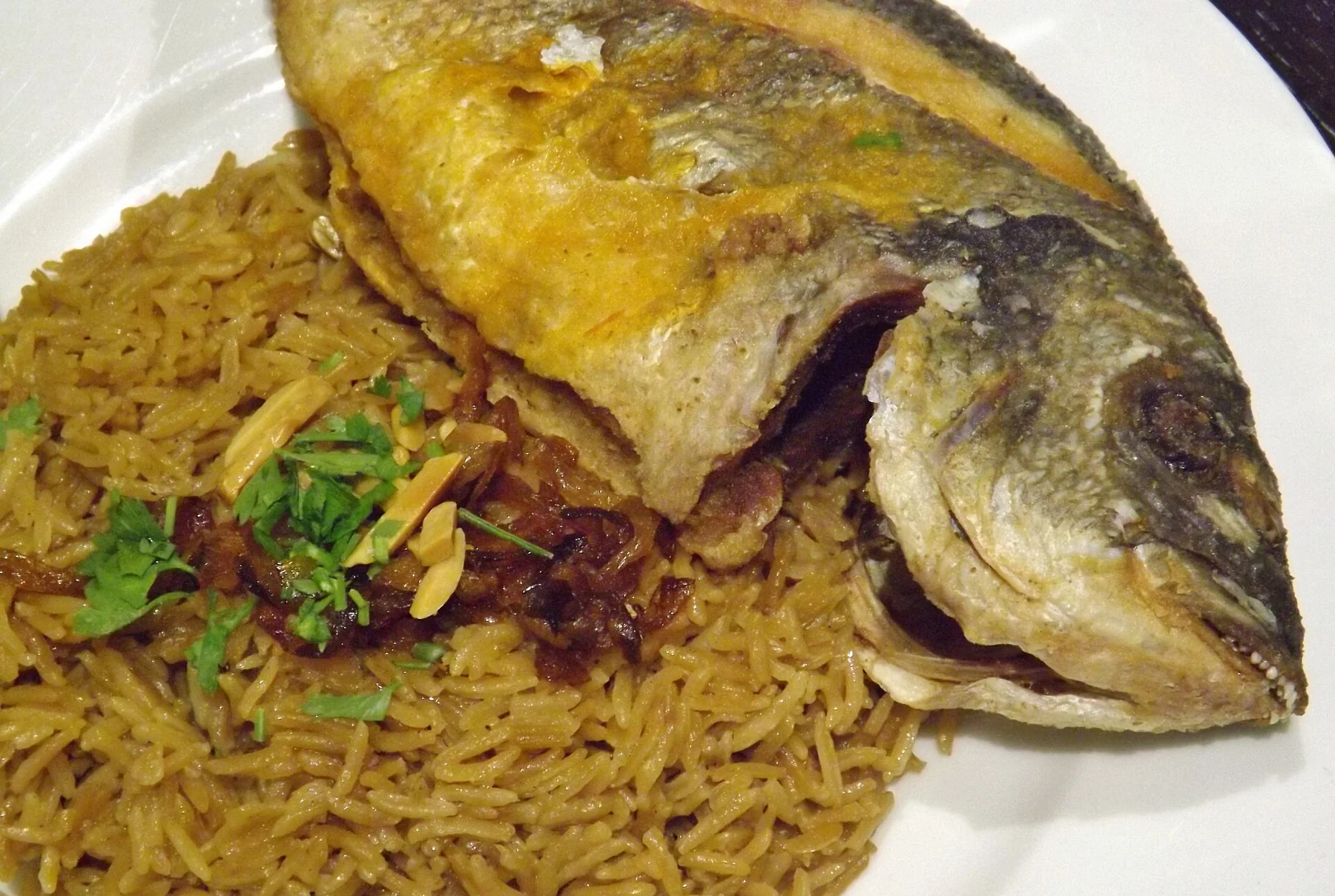 A whole fried fish on a bed of brown, onion-dark rice scattered with herbs.