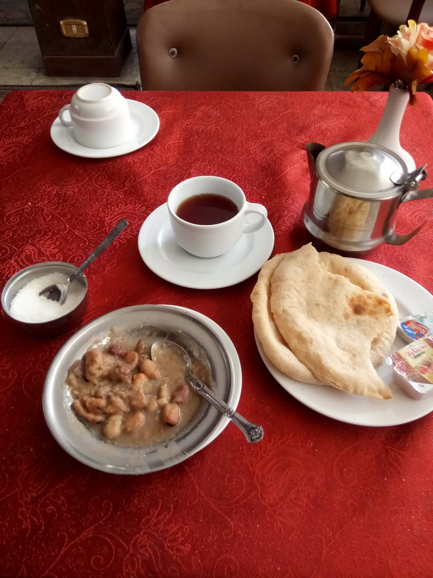 On a red tablecloth, a metal bowl of whole beans with a spoon, flatbread on a plate, and a cup of tea.