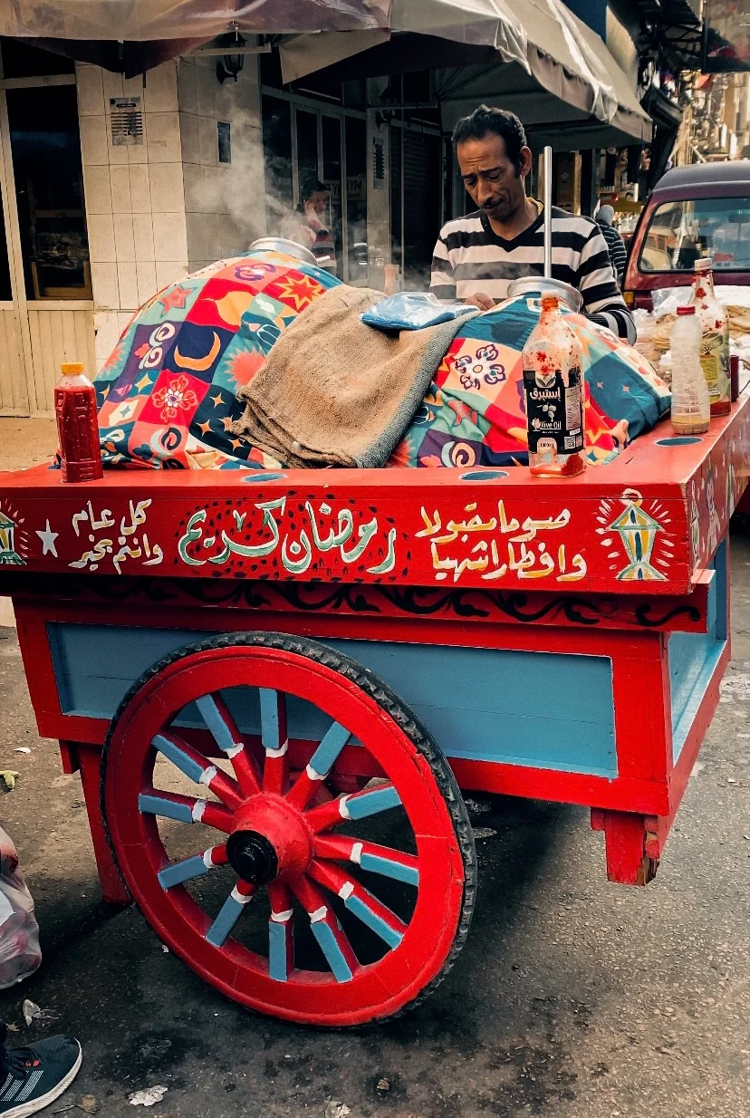 A red wooden street cart on a large red wheel. Its load is wrapped in patterned cloth and sacking, steam rises, and a vendor stands behind it.