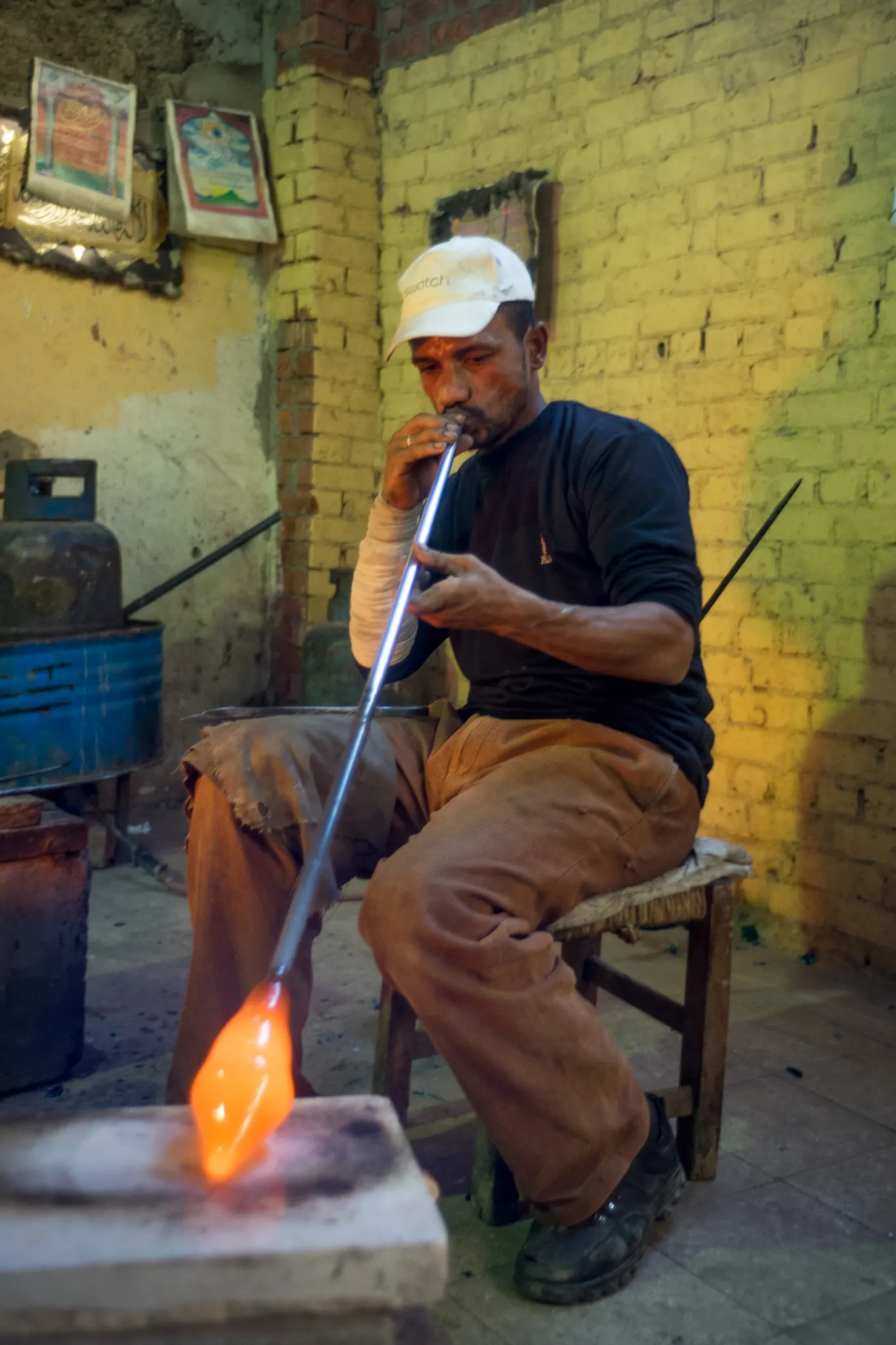 A glassblower in Cairo seated at his bench, blowing through a long metal pipe, in a photograph from 2016.
