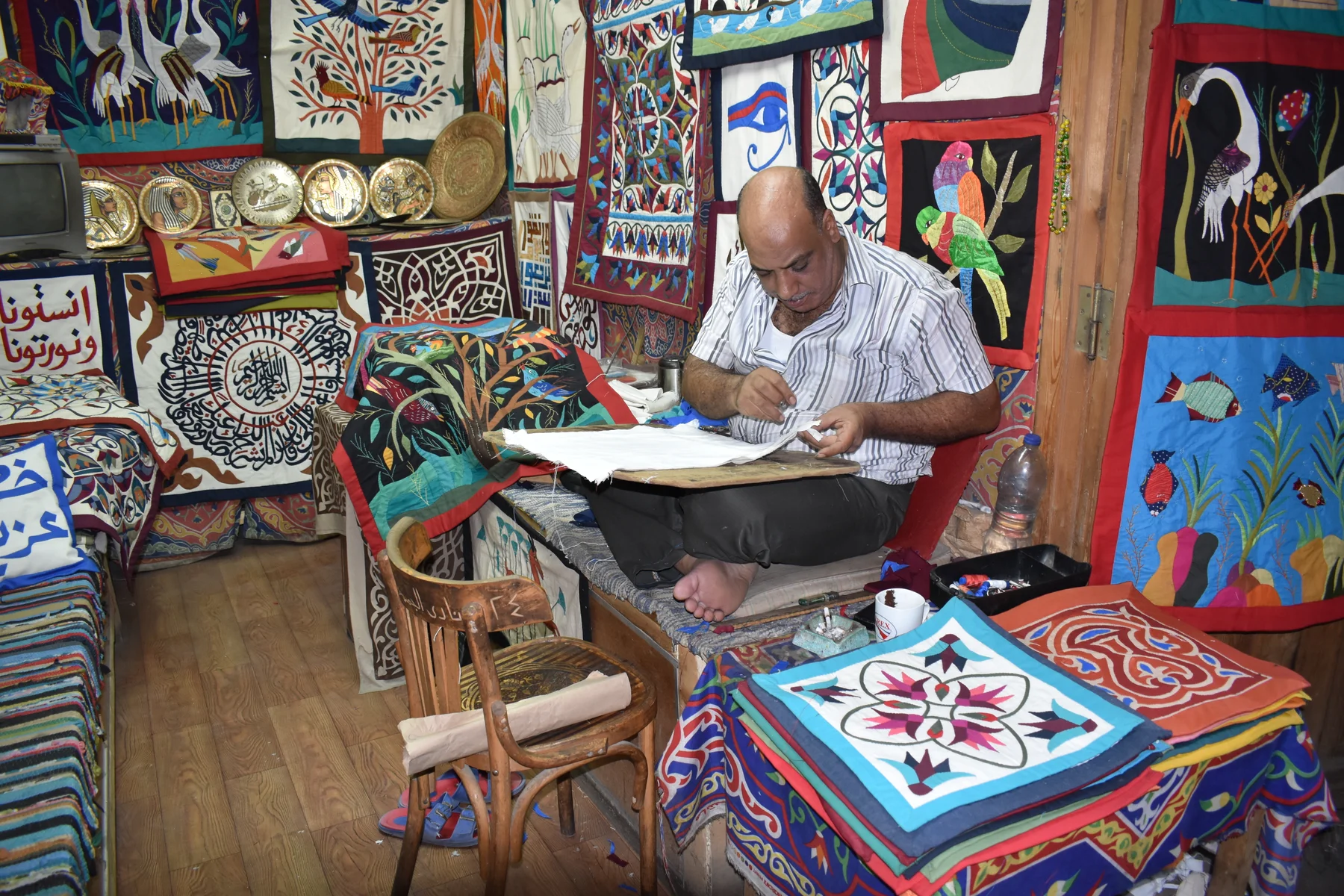 A seated tentmaker working with white cloth across his lap, surrounded by hanging appliqué panels, in a photograph from 2017.