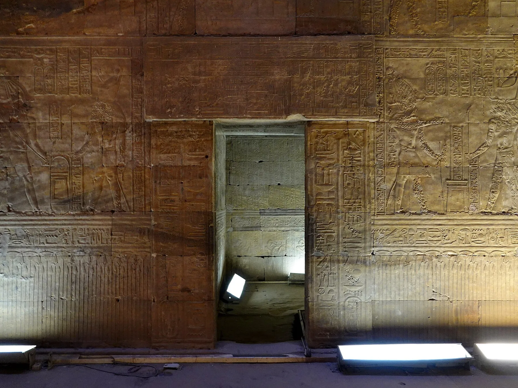 A narrow doorway in a carved temple wall, lit from the floor. Through it, the far wall of a small room is covered in columns of inscription.