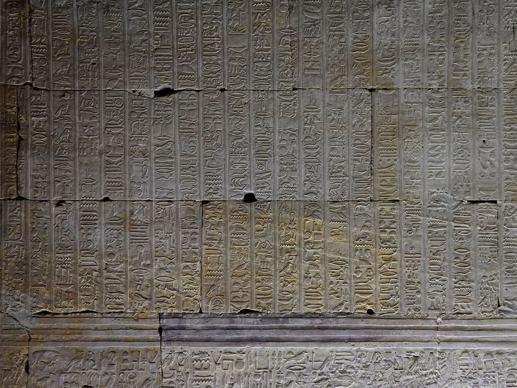 A stone wall covered in columns of carved hieroglyphic signs, broken by cracks and round holes, with a band of larger signs along the bottom.