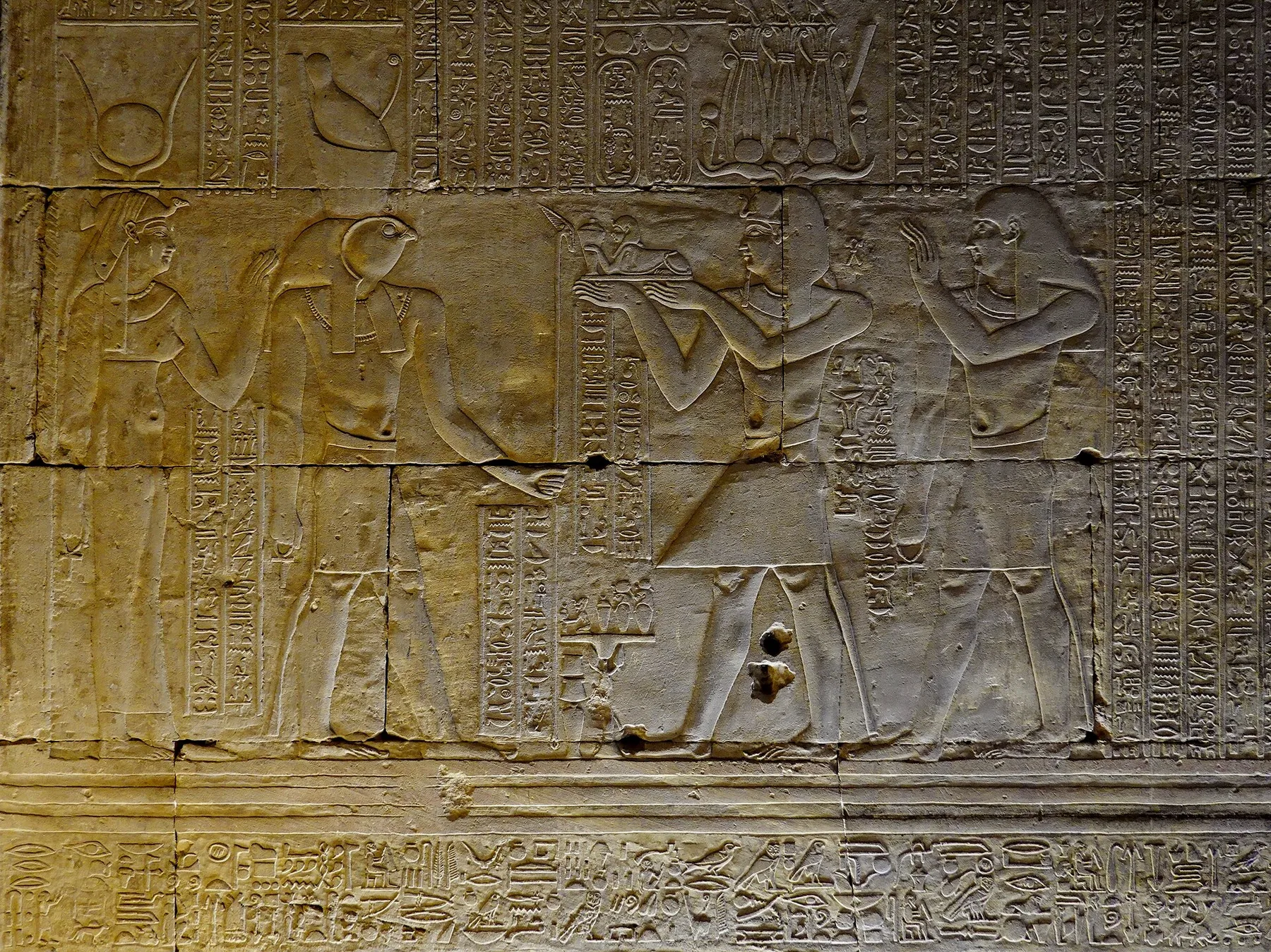 A carved relief: a crowned king holds out a small tray towards a falcon-headed god and a goddess wearing horns and a sun disc; a second figure behind the king raises both hands.