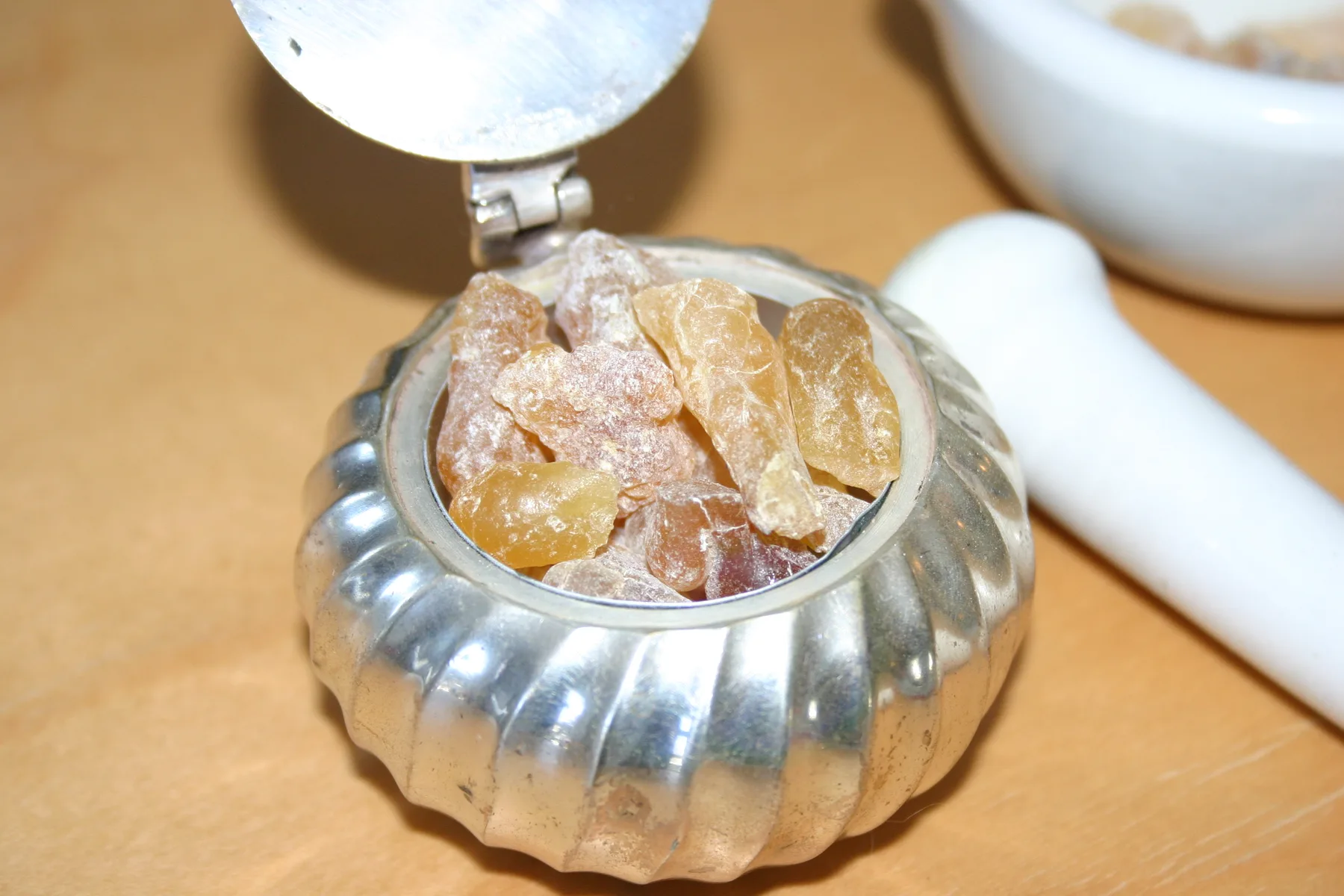 Translucent amber lumps of frankincense in a fluted silver dish, a white mortar and pestle behind.