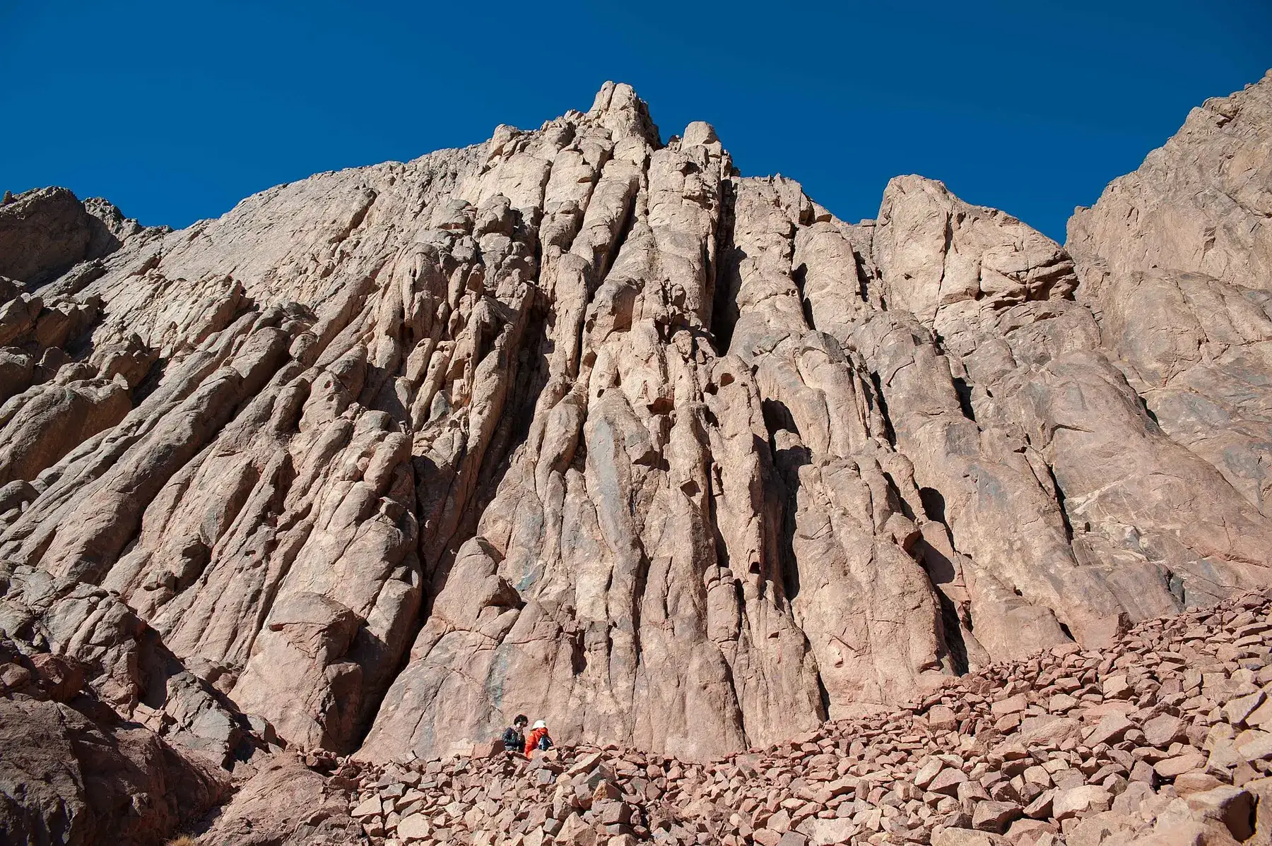 Granite cliffs beside a trail on Mount Catherine in South Sinai, with two walkers at their foot.