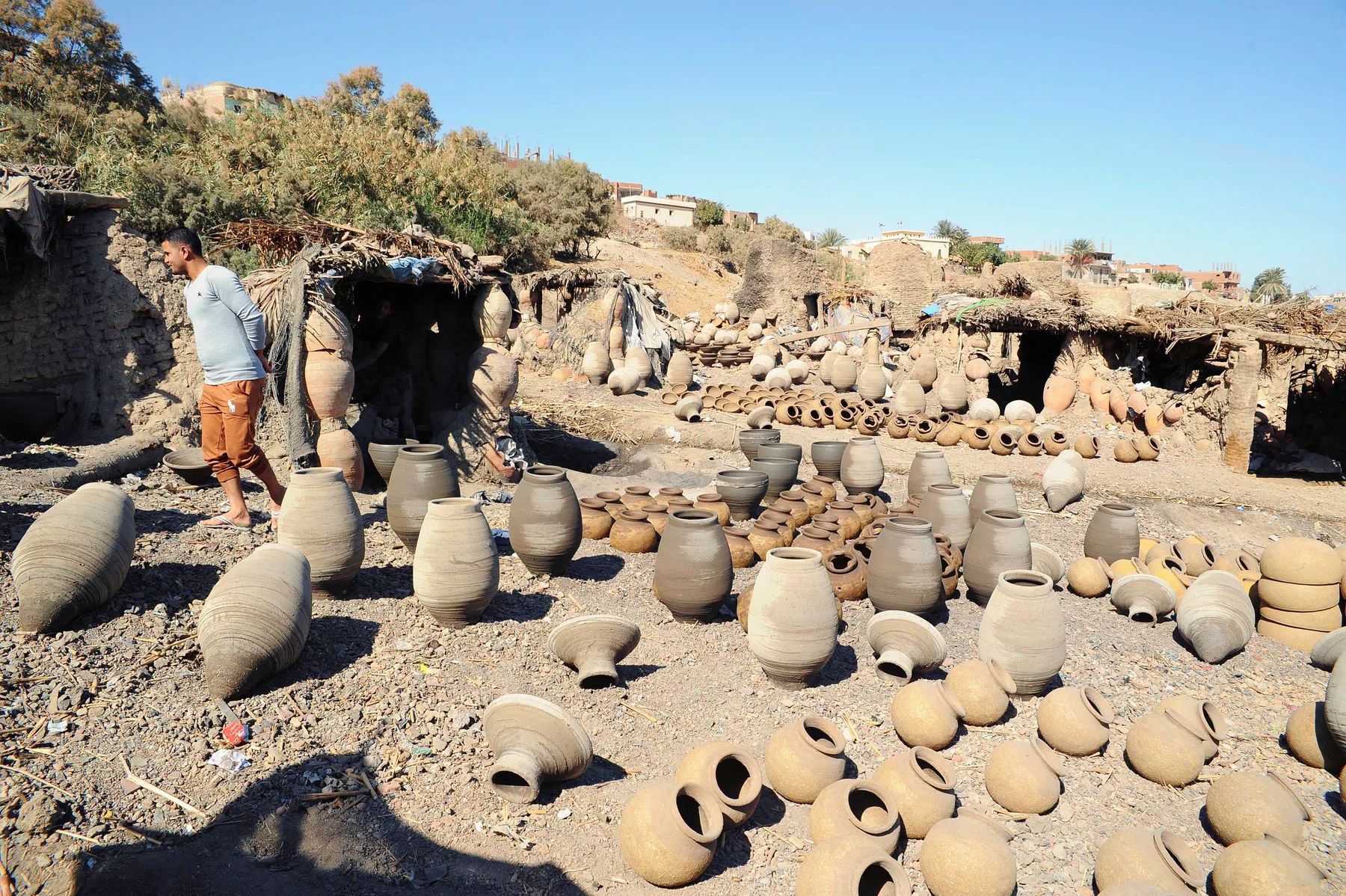 Unglazed jars drying on open ground outside reed-roofed work sheds. Tall grey jars with ringed sides stand in rows; smaller, paler round pots lie in the foreground. A man stands at the left.