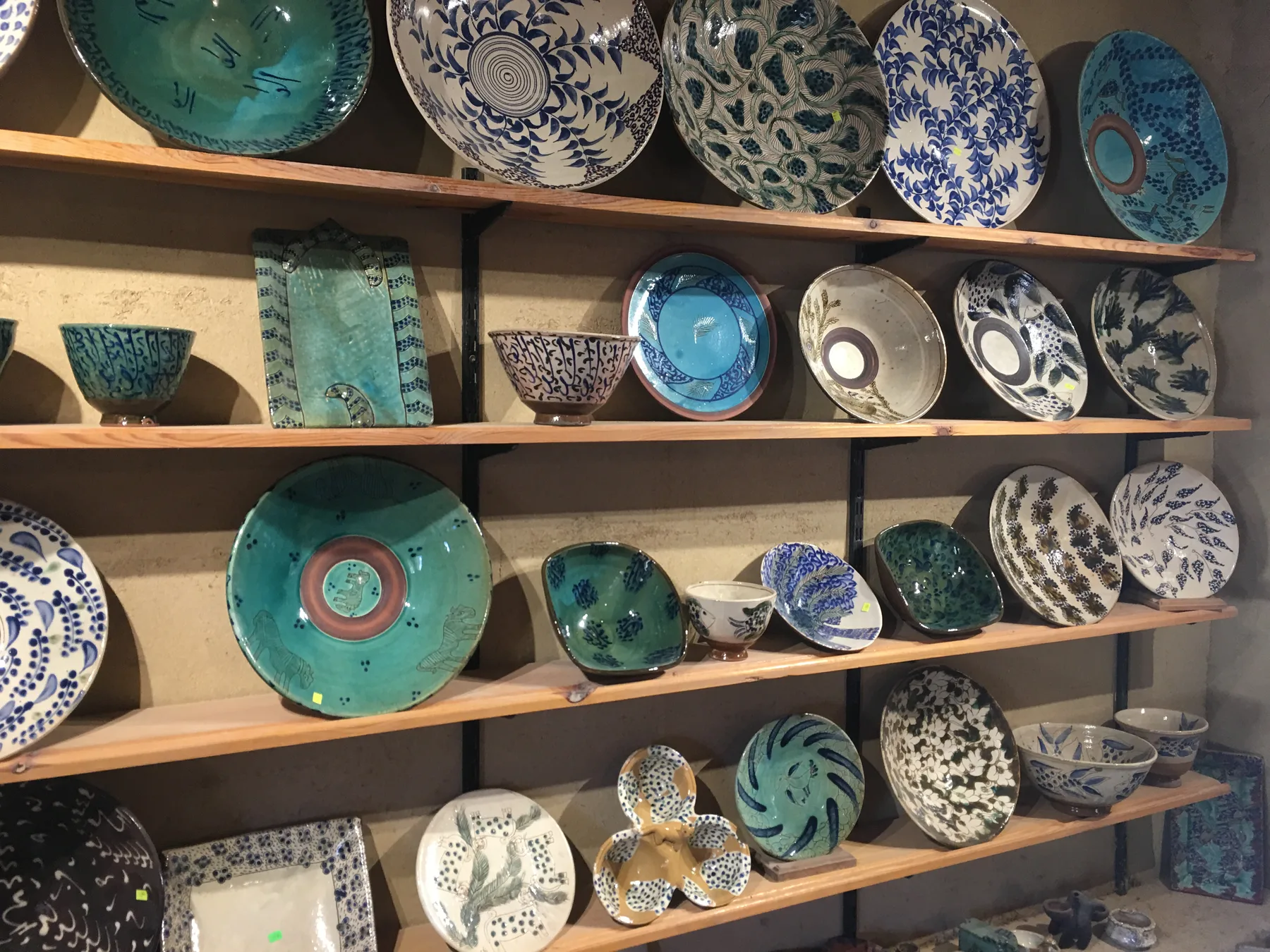 Glazed bowls and plates on wooden shelves: turquoise, cream and white grounds, painted in blue and dark green with leaves, spots, letter-like marks and animals.