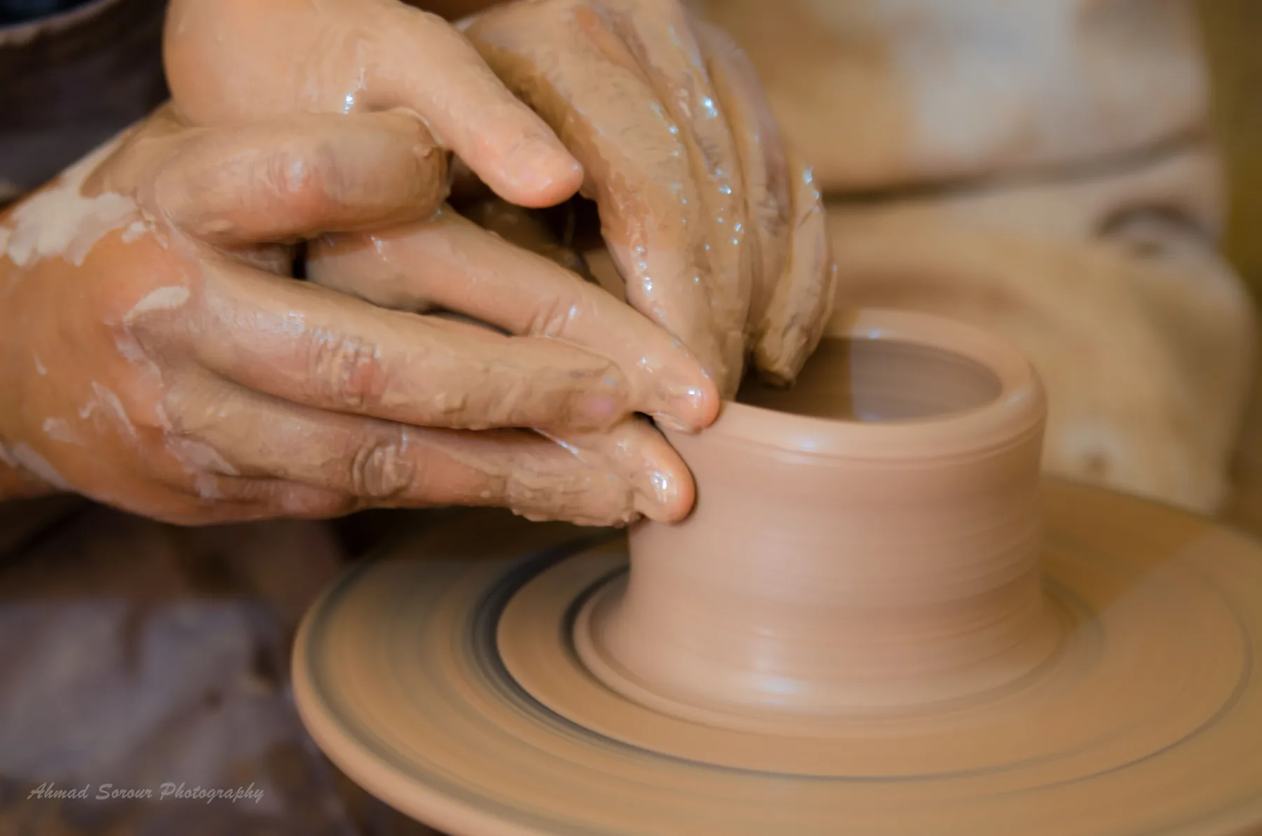 Two clay-covered hands shaping a small cylinder of clay on a turning wheel head.
