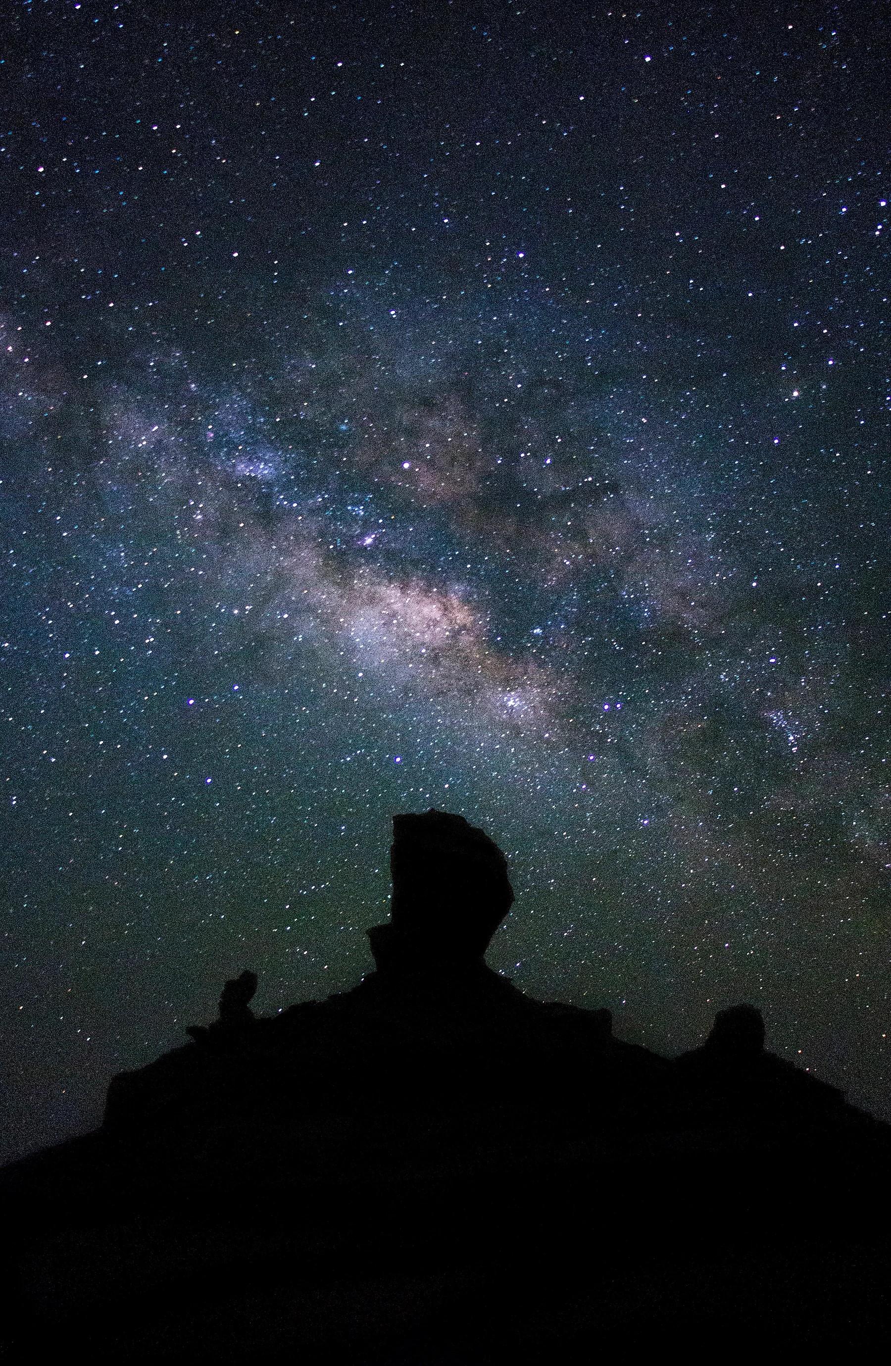 The Milky Way, a broad pale band of stars with darker lanes, crossing the sky above the black silhouette of a rock formation.