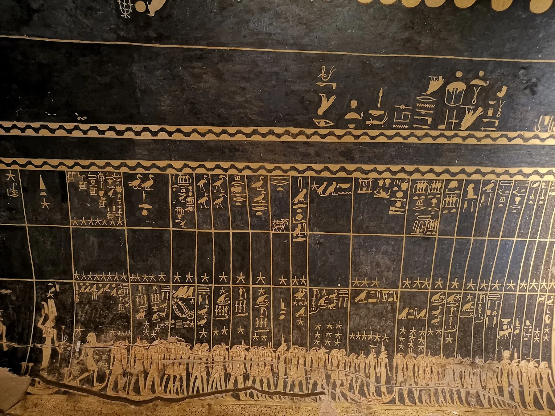 Part of a tomb ceiling painted pale gold on black: columns of text with rows of stars above a procession of figures.