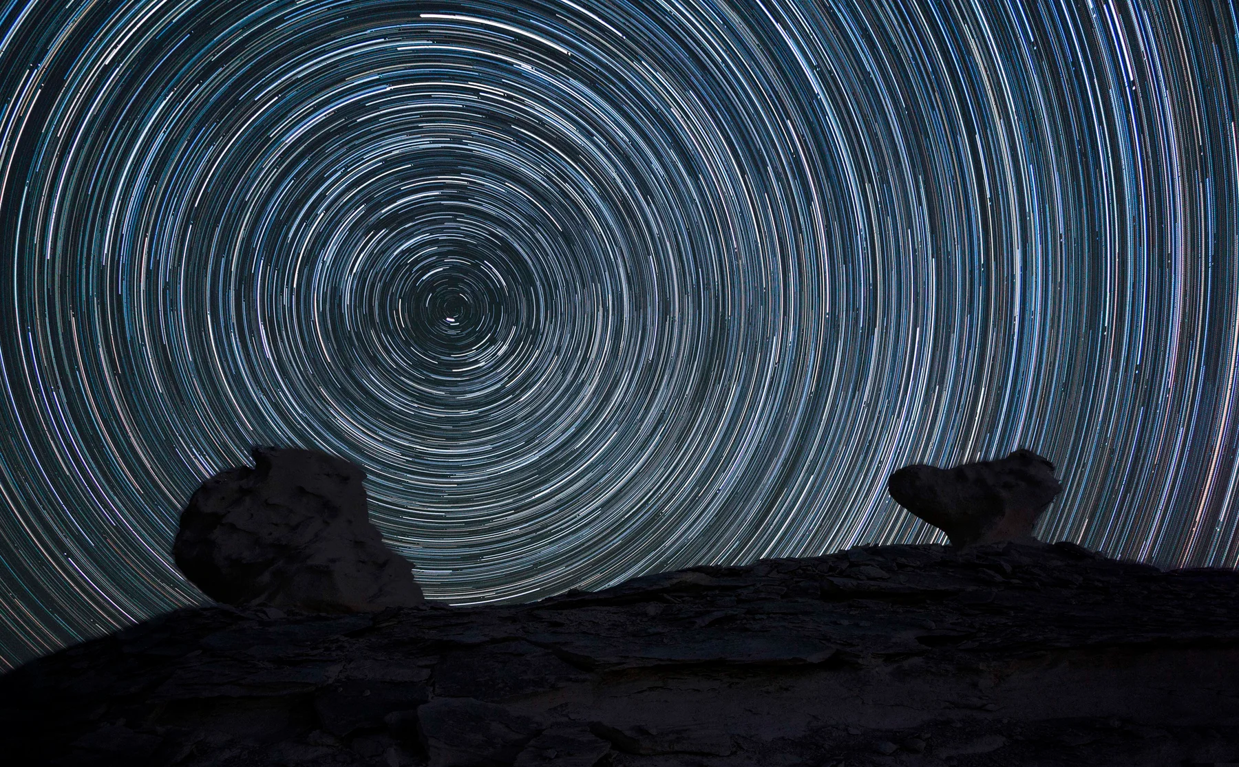 A long-exposure photograph: concentric arcs of starlight share one centre above two dark rock formations.