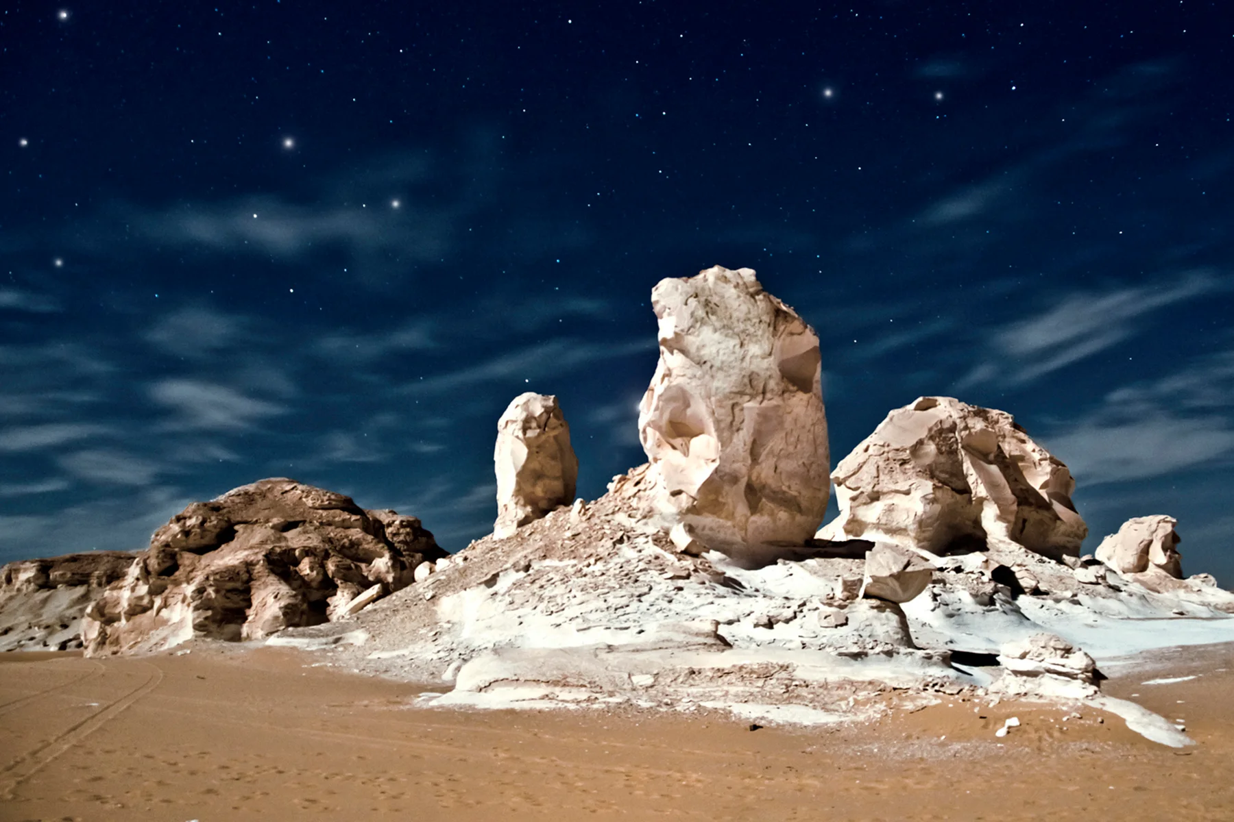Pale chalk formations lit against a deep blue night sky full of stars, with sand in the foreground.