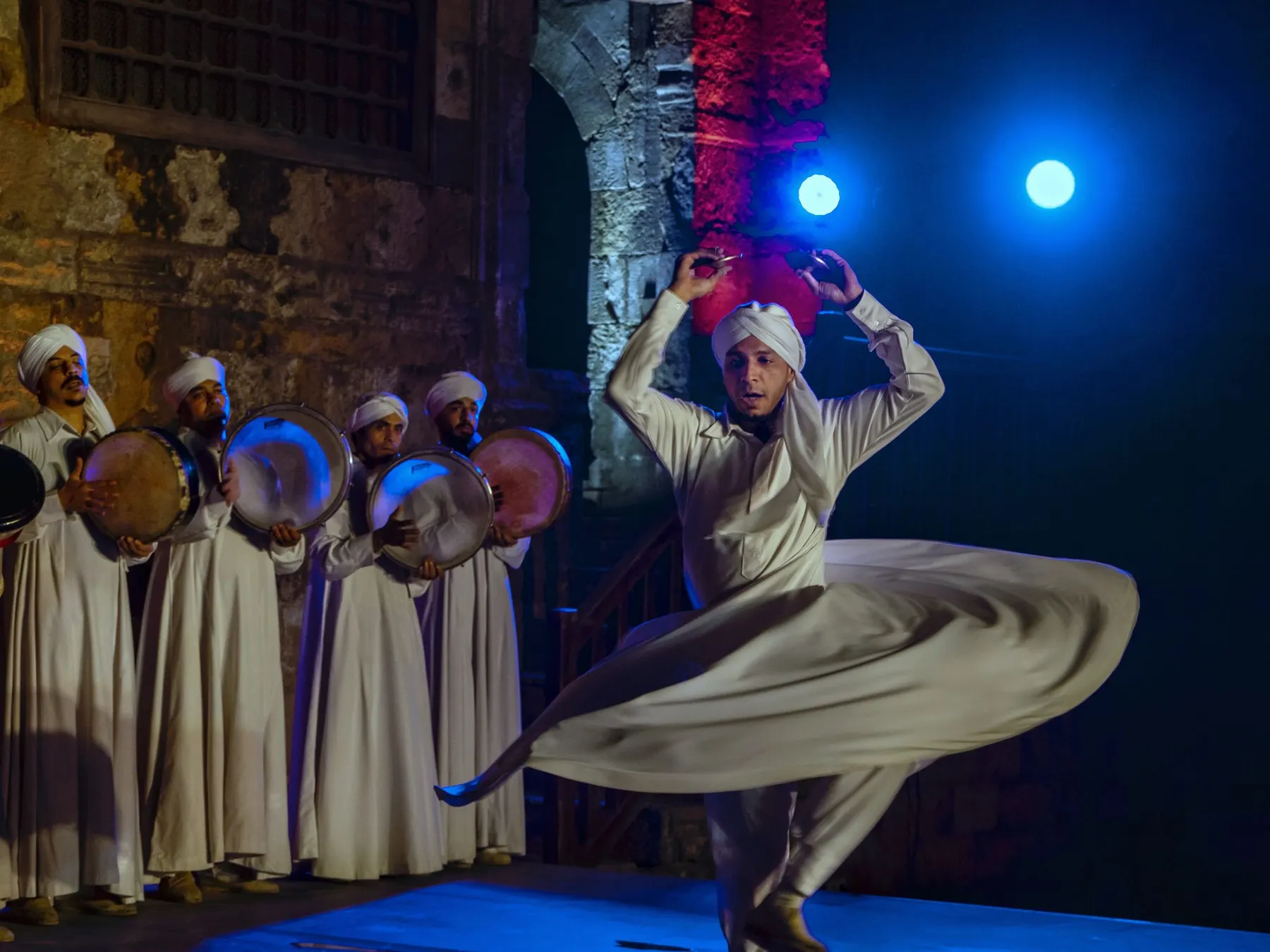 A Tanoura dancer spinning in a white robe while drummers play behind him, in Cairo in 2021.