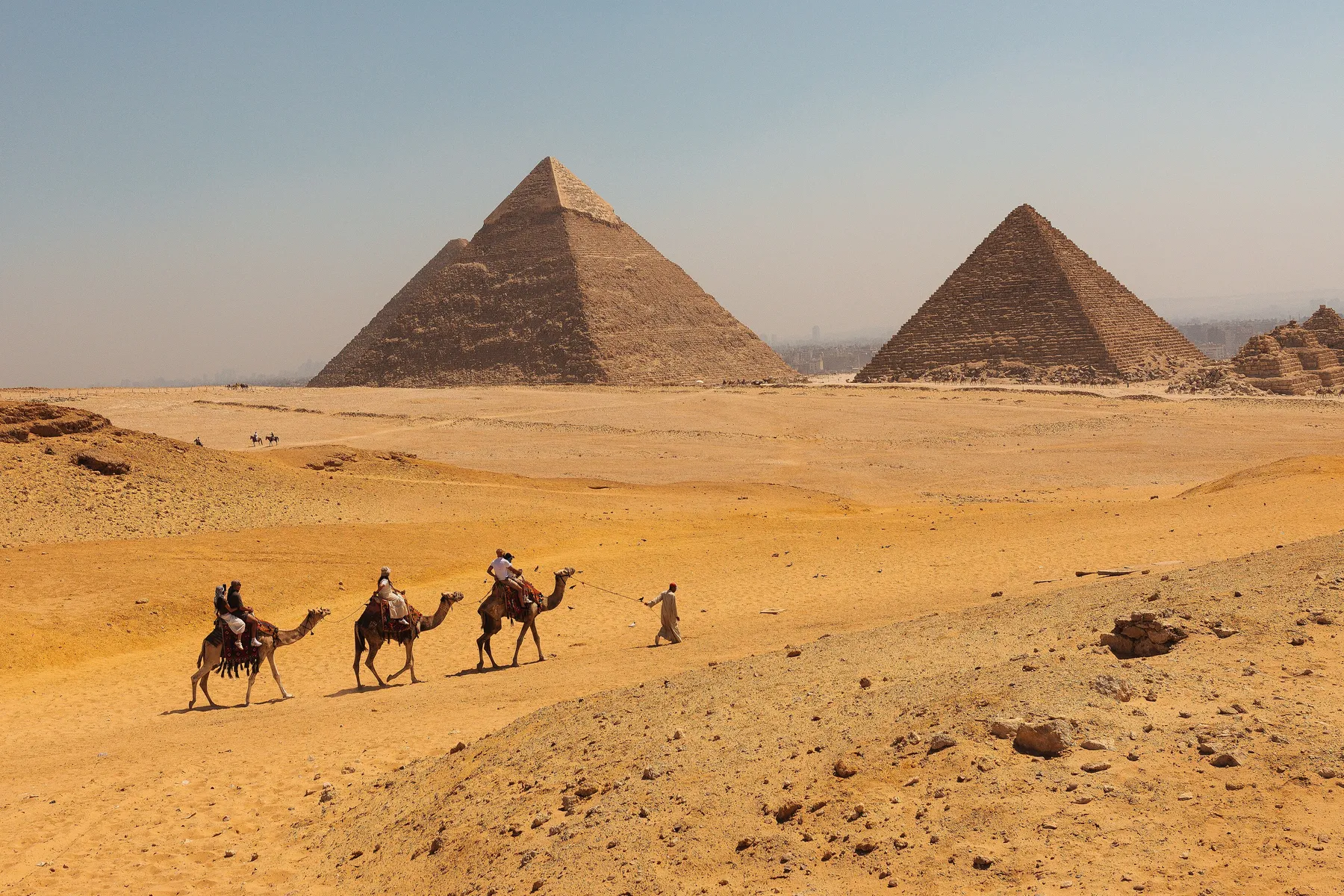 Three riders on camels crossing the sand below the pyramids of Giza.